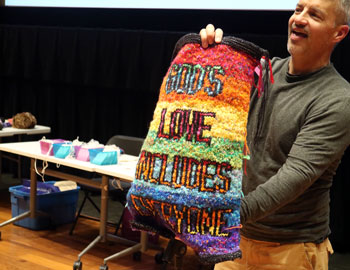A man holds out a hand knitted rainbow pulpit fall that reads 'God's love includes everyone'