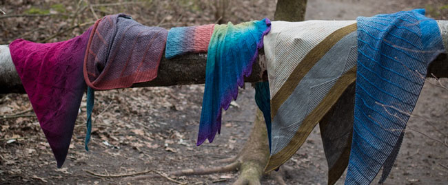 A group of colourful knit scarves hung along a log in a forest.